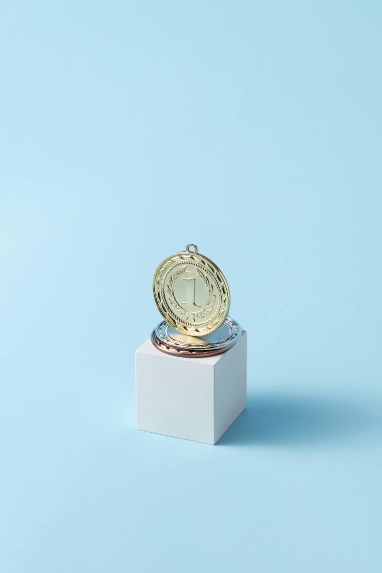 A gold medal displayed on a cube against a minimalist light blue background, showcasing achievement.